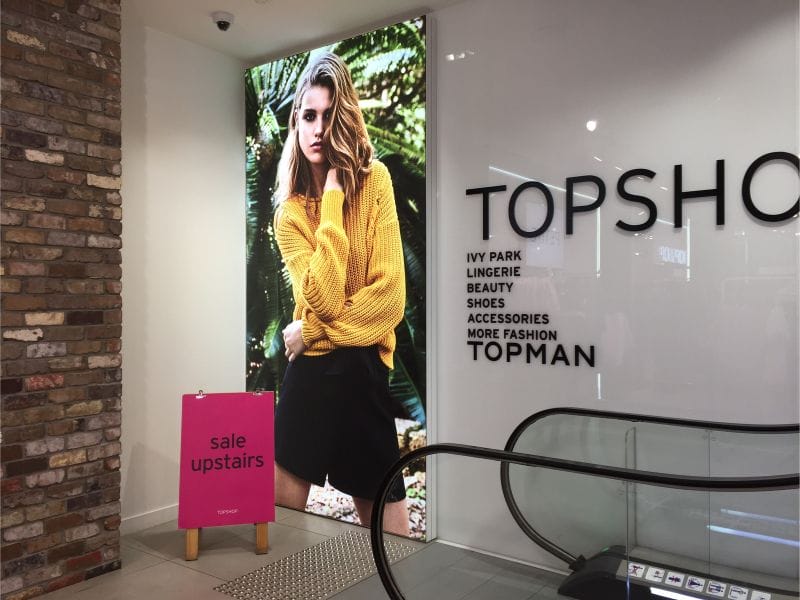 Illuminated portrait orientation of wall-mounted Brandframe at fashion store escalators - Displays2Go